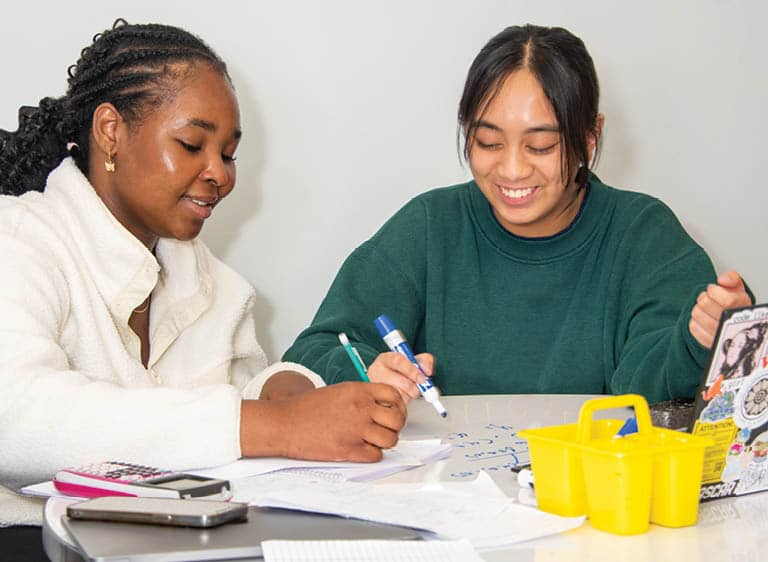 Two FLCC students writing on a whiteboard together, with a laptop and water bottle nearby.