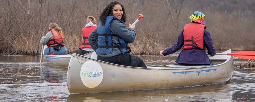 Four students canoeing near a shoreline on a brisk autumn day. One rower looks back over her shoulder as the other three look onward.