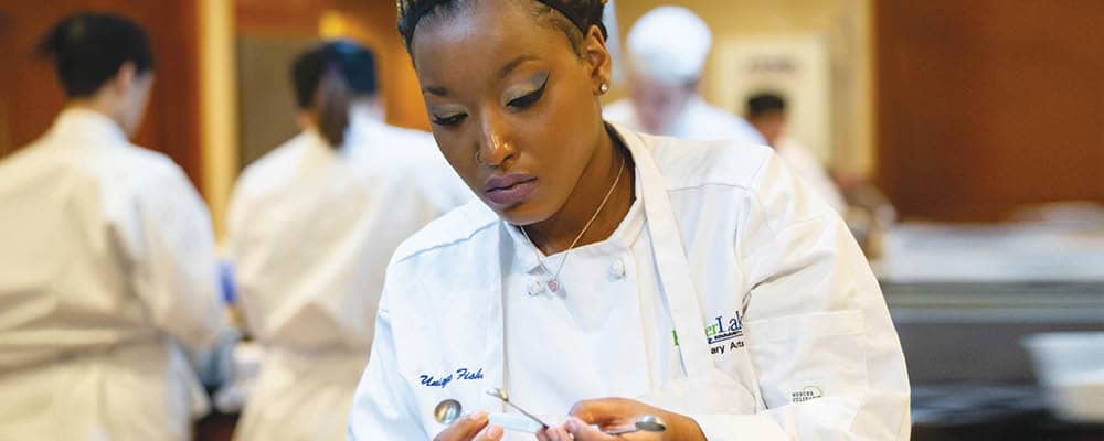 A student in a white chef's jacket and apron. She inspects a set of measuring spoons as other students prepare food in the background.