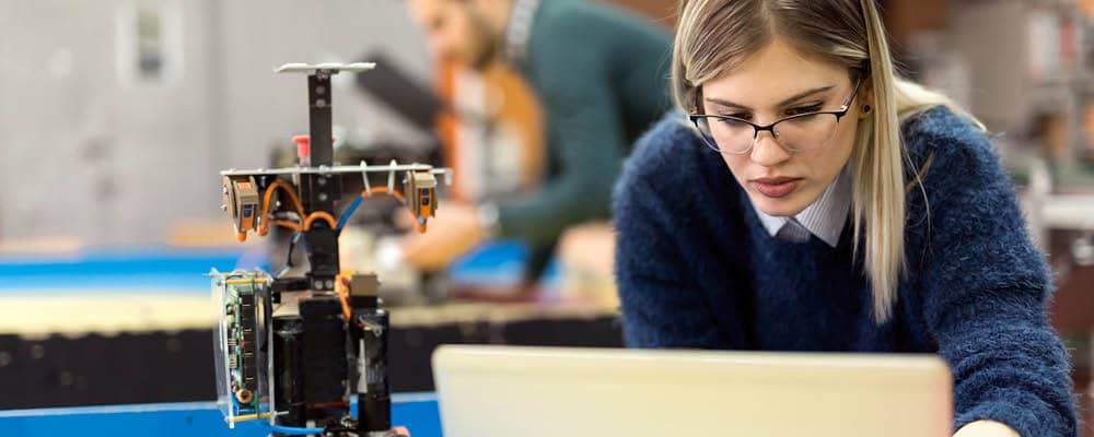 A Smart Systems Technologies student working on a laptop, programming a nearby robotic device.